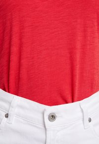 Rød bomulds T-shirt parret med hvide denim bukser. T-shirten har en glat, let struktureret overflade; bukserne har en sølvknap og bæltestropper.