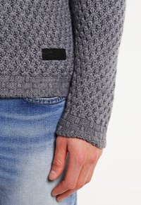 Ruka odpočívající vedle světle modrých džín s texturovaným šedým pleteným rukávem svetrku, na kterém je malý černý obdélníkový štítek.