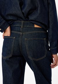 Pantalones vaqueros de mezclilla oscura con un diseño ajustado, costuras naranjas en contraste y dos bolsillos traseros, con una etiqueta de cuero marrón claro en la cintura.