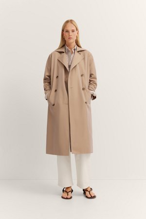 Beige double-breasted trench coat with large pockets, wide collar, long sleeves, and a smooth texture, worn over a striped shirt and white pants.