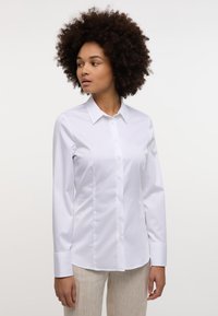 Camicia bianca con colletto a punta, maniche lunghe e una texture liscia e lucida. Presenta bottoni frontali e una vestibilità sartoriale.