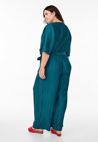 Turchinfarbener, plissierter Jumpsuit mit tailliertem Gürtel, kurzen Ärmeln und weitem Bein, kombiniert mit roten Sandalen. Rückansicht, die Textur und gestreiftes Muster zeigt.