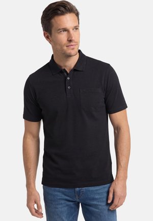 Mann trägt ein schwarzes kurzärmeliges Poloshirt mit einer Brusttasche und blaue Jeans, blickt leicht nach rechts vor einem einfarbigen Hintergrund.