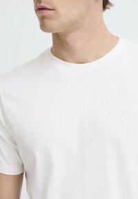 Camiseta de algodón blanca con escote redondo, que presenta un sutil logo bordado en la esquina superior izquierda. Manga corta y textura suave.