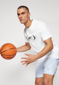 Hvit kortermet skjorte med svart logo, kombinert med lyseblå shorts med perforert tekstur og hvite mønstrede detaljer. Holder en basketball.