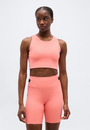 Femeie atletă purtând un top scurt fără mâneci de culoare roz coral și pantaloni scurți biker cu talie înaltă, pe un fundal simplu.