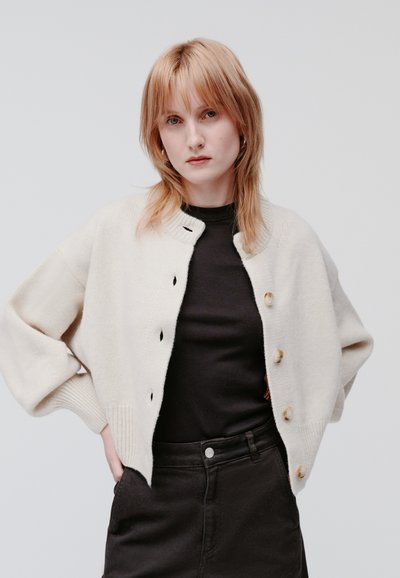 Crème-kleurige cardigan met een ronde hals, knopen sluiting, geribbelde manchetten en verlaagde schouders, gedragen over een zwarte top en rok.