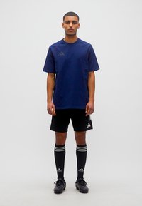 Άνδρας που στέκεται με το πρόσωπο προς τα εμπρός φορώντας μπλε ναυτικό αθλητικό μπλουζάκι, μαύρο σορτς Adidas, μαύρες κάλτσες Adidas μέχρι το γόνατο και μαύρα ποδοσφαιρικά παπούτσια με τακούνια.