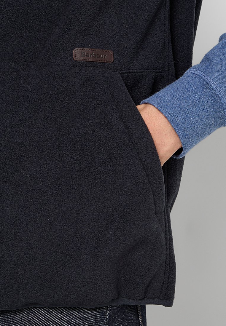 Pull en polaire noir avec une poche avant. Il comporte une étiquette en cuir marron estampillée "Barbour". Une chemise bleue à manches longues est visible en dessous.