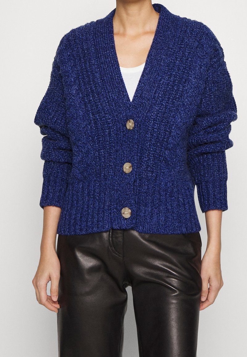 Blå stickad cardigan med djup V-ringning, ribbade muddar och tre stora knappar. Bärs över en vit topp och matchas med svarta läderbyxor.