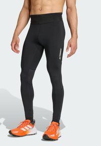 Black athletic leggings with a fitted design, textured mesh panels, and a wide waistband. A white logo is visible on the thigh. Bright orange running shoes complete the outfit.