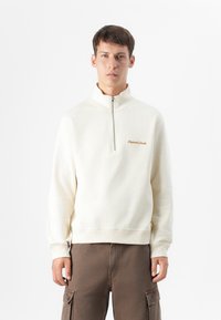 Ung mand står vendt fremad, iført en cremet zip-up sweatshirt med et lille broderet logo, parret med brune cargo bukser.