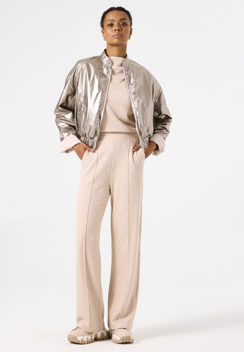 Metallic silver bomber jacket, beige sweater, and high-waisted wide-leg beige pants. Chunky beige sneakers with wavy sole.