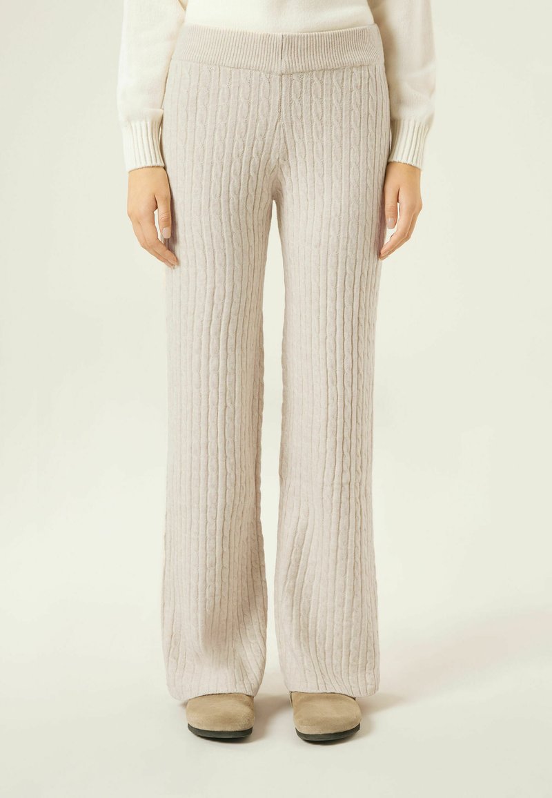 Jasnobeżowe spodnie o szerokich nogawkach wykonane z dzianiny warkoczowej, z elastycznym paskiem w talii. Tkanina ma miękką, fakturowaną strukturę. Podkreślone przez neutralne obuwie.