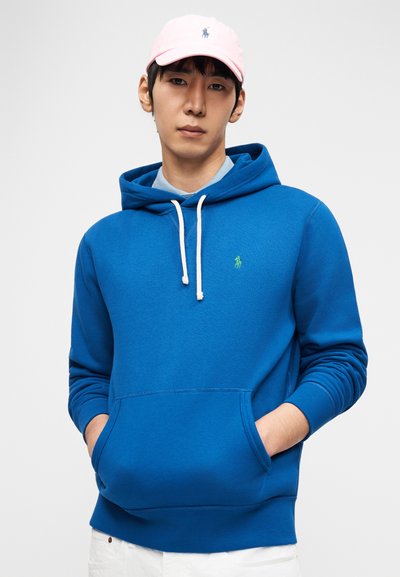 Νέος άνδρας με μπλε φούτερ με κουκούλα και λευκά κορδόνια, φοράει ροζ καπέλο, με τα χέρια στην τσέπη του φούτερ, μπροστά σε απλό ανοιχτόχρωμο φόντο.