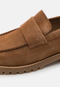 Chaussure mocassin en daim marron avec un bout arrondi, des détails cousus, une semelle en caoutchouc et un logo embossé sur le côté près de la bride.