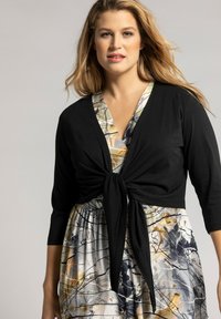Cardigan noir noué sur une robe à motifs multicolores comportant des designs abstraits, avec des manches trois-quarts et un décolleté en V.