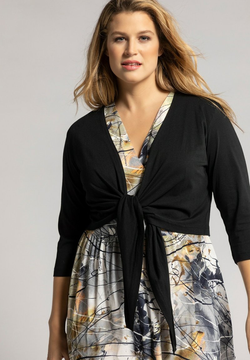 Cardigan noir noué sur une robe à motifs multicolores comportant des designs abstraits, avec des manches trois-quarts et un décolleté en V.