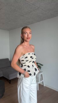 Trägersloses Oberteil in Creme mit schwarzen abstrakten Punkten, kombiniert mit weißen hoch taillierten Hosen. Das Oberteil hat Taschen und einen strukturierten Stoff. Einfaches Design.