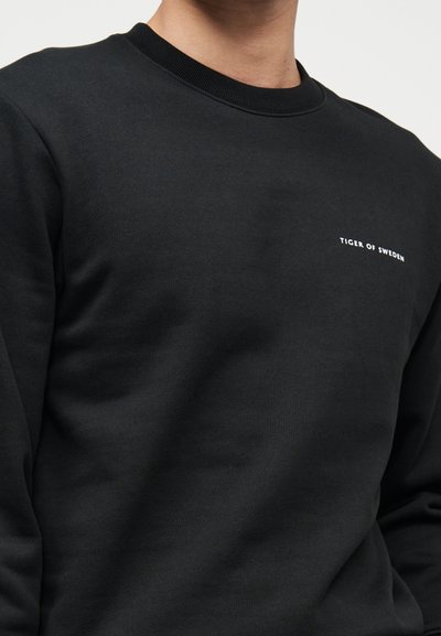 Μαύρο φούτερ με στρογγυλή λαιμόκοψη, με μικρό λευκό λογότυπο κειμένου "Tiger of Sweden" στο αριστερό στήθος, φορεμένο από ένα άτομο από τον λαιμό μέχρι τον κορμό.