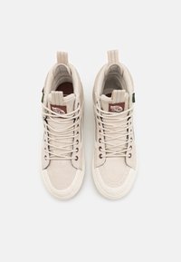 Pari vaaleanbeigejä high-top Vans -tenniskenkiä, joissa on valkoiset nauhat, vetoanalyysit ja ruskeat "Off The Wall" -logo-patchit kielissä.