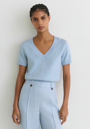 Kobieta z warkoczem ma na sobie jasnoniebieski sweter z krótkim rękawem i dekoltem w serek oraz spodnie z wysokim stanem na tle gładkiej białej ściany.