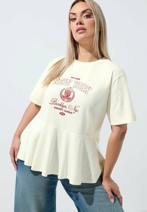 Blond kvinde iført en cremefarvet T-shirt med flæset kant og rød New York varsity-print og blå jeans, poserer mod en ensfarvet lys baggrund.