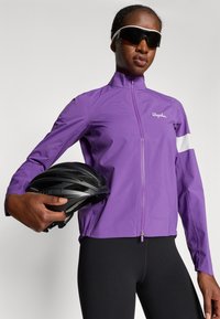 Paarse fietsjack met een hoge kraag, ritssluiting aan de voorkant en witte accenten. Model houdt een zwarte helm vast. Zwarte aansluitende broek.