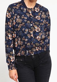 Marinblå blommig bomberjacka med en lätt åtsittande midja, prydd med rosa och gula blommönster samt svarta knappar, matchad med svarta jeans.