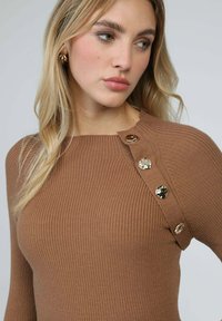 Pull côtelé marron avec un col rond et des boutons décoratifs dorés sur l'épaule gauche, présentant une texture lisse.