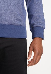 Main droite d'une personne portant une chemise bleue à manches longues et un pantalon noir sur un fond blanc uni.