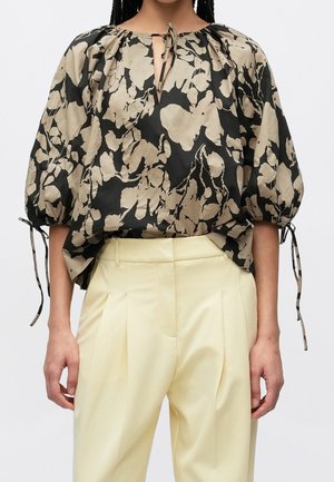 Femme portant un chemisier ample noir et beige à motif floral avec des manches bouffantes et un pantalon plissé taille haute jaune clair.