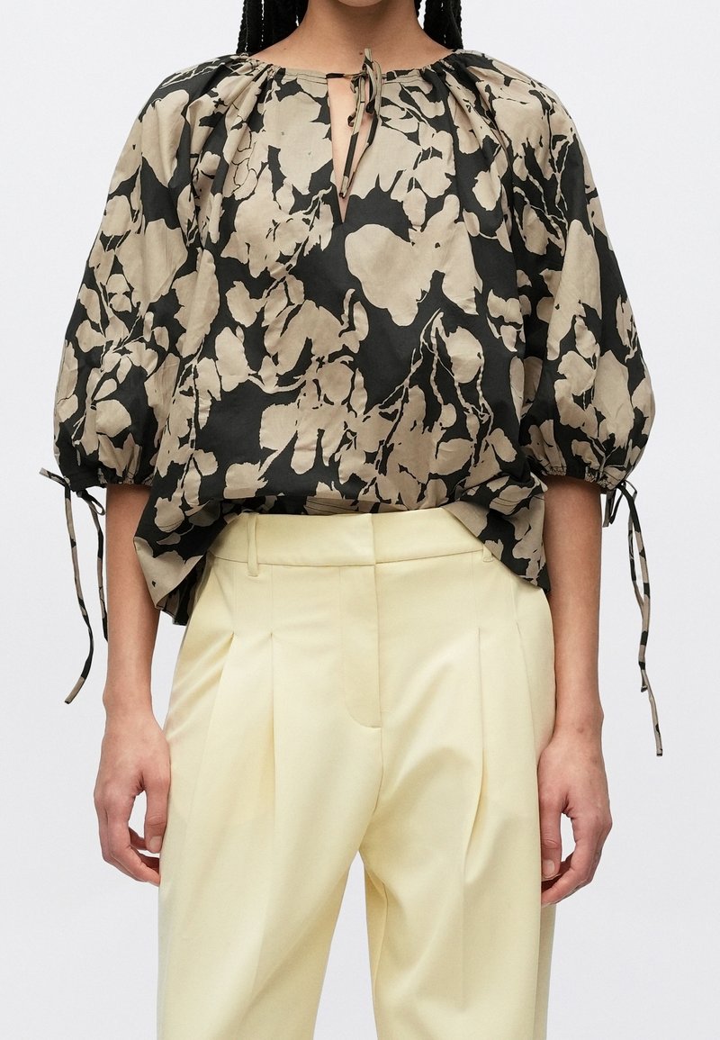 Femme portant un chemisier ample noir et beige à motif floral avec des manches bouffantes et un pantalon plissé taille haute jaune clair.