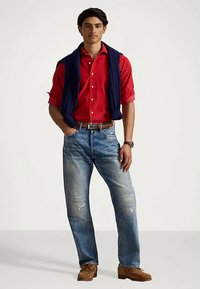 Raudona marškinėliai su sagomis, tamsiai mėlynas megztas liemenė, išblukusios mėlynos džinsai su dėvėjimo žymėmis, rudas diržas ir zomšos mokasinai. Švarus fonas.