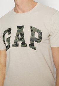 Camiseta de algodón beige claro con un gran logotipo de "GAP" en camuflaje en tonos verdes y negros. Diseño clásico de cuello redondo con mangas cortas.
