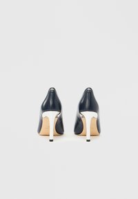 Marineblå høye helsko i skinn med spisse tupper og sølvfargede stilett-hæler. Jevn tekstur, minimalistisk design med en subtil logo på hælen.