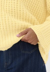 Gros plan sur une main ajustant un pull jaune côtelé sur un jean bleu foncé avec des coutures de poche visibles.