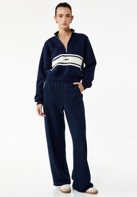 Sudadera azul marino con cierre y rayas blancas, acompañada de pantalones anchos azul marino a juego. El material parece suave y grueso.