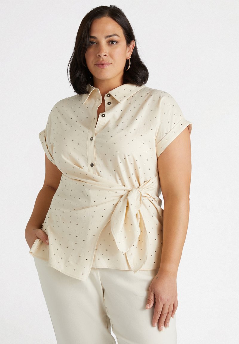 Camicia crema con bottoni, maniche corte arrotolate, caratterizzata da un nodo in vita e un delicato motivo a pois in toni metallici.