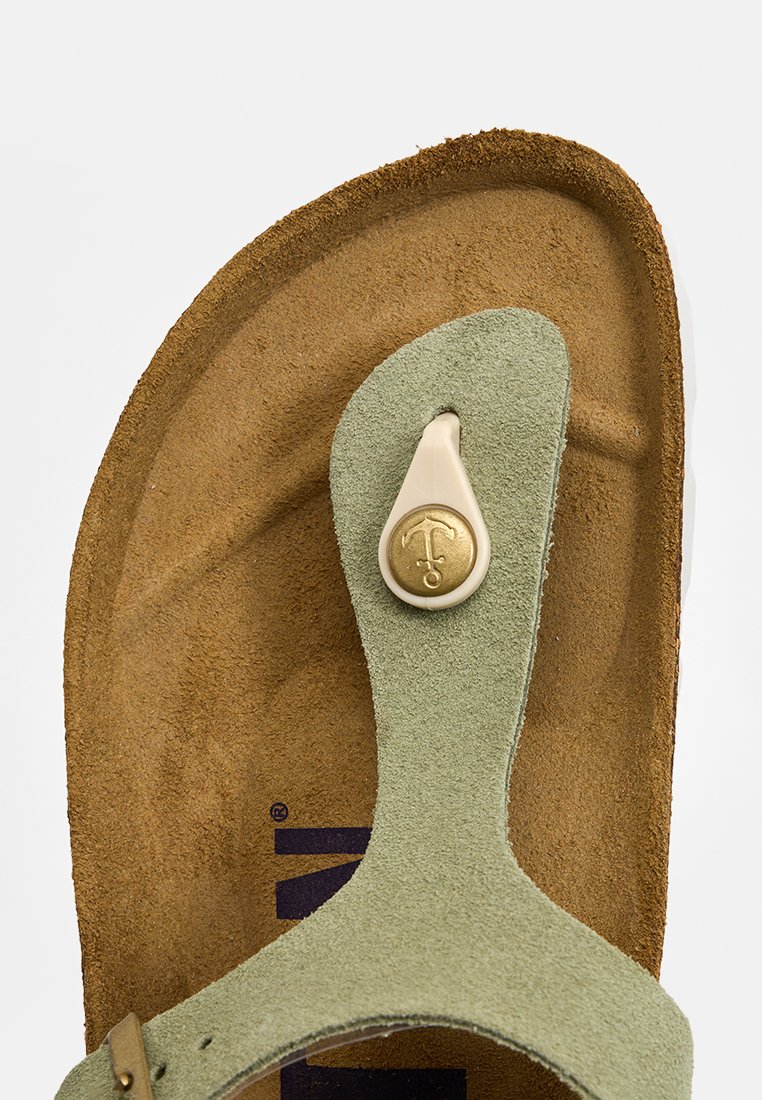 Nahaufnahme eines braunen Fußbettsandals mit hellgrünem Wildlederriemen und rundem, beigem Kunststoffzehensteg mit goldenem Ankeremblem.