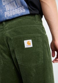 Zelené manšestrové kalhoty s texturovaným povrchem, na zadní kapse s logem značky Carhartt a viditelnými švy.