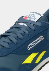 Mėlyni zomšiniai sportbačiai su geltonais akcentais, plokščiomis raištelėmis ir Reebok logotipu. Turi minkštą tekstūrą ir baltą liežuvėlį su spaustu raštu.