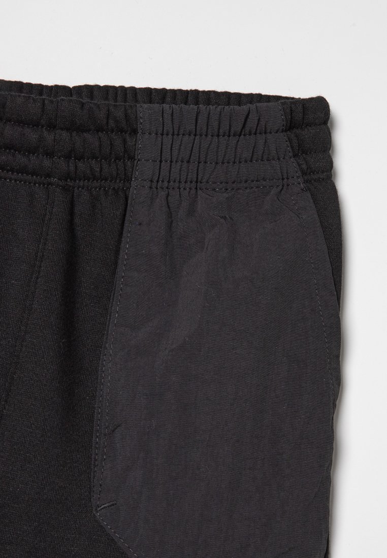 Pantalones de chándal negros con cintura elástica, textura suave y un bolsillo lateral con un panel en contraste. Diseño simple y funcional.
