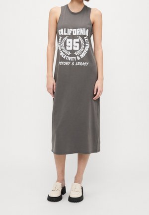 Person iført grå midi-kjole uden ærmer med teksten "California 95 Freedom Creativity & Individuality History & Legacy" og cremefarvede loafers.