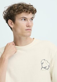 Cremefarvet bomulds t-shirt med rund halsudskæring og korte ærmer. Har et grafisk billede af en bomuldsplante på venstre brystområde.
