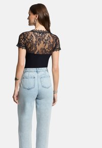 Musta bodysuit, jossa on pitsinen selkä ja lyhyet hihat, yhdistettynä vaaleansinisiin korkean vyötärön farkkuihin. Pitsissä on kukkakuvioita ja sahalaitaiset reunat.