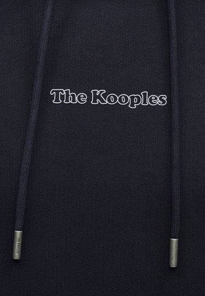 Gros plan sur un sweat à capuche sombre avec le logo "The Kooples" imprimé en blanc et des cordons assortis avec des embouts en métal.