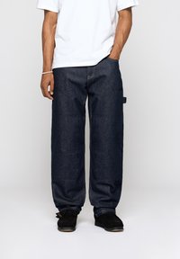 Tummansiniset farkut, joissa on väljä malli, suora lahjetyyli ja kontrastiväriset tikkaukset. Yhdistetty valkoiseen t-paitaan ja mustiin kenkiin.