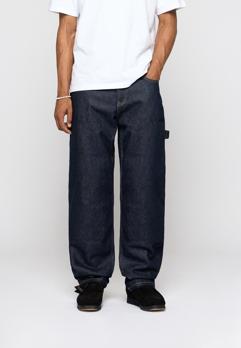 Tummansiniset farkut, joissa on väljä malli, suora lahjetyyli ja kontrastiväriset tikkaukset. Yhdistetty valkoiseen t-paitaan ja mustiin kenkiin.