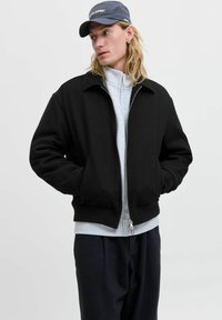 Zwarte bomberjack met een kraag, geribde manchetten en een rits. Gekleed over een grijze sweatshirt, gecombineerd met donkere broeken en een grijze pet.